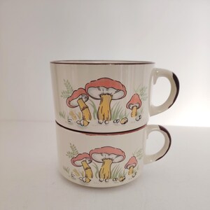 May include: Two stacked cream-coloured ceramic mugs featuring a repeating illustration of three mushrooms with pinkish-red caps, yellow stems, and green grass accents. Each mug has a dark brown rim and a matching dark brown handle on the right side.