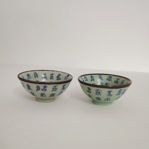 May include: Two small, light green ceramic bowls with a dark brown rim and blue Chinese characters. The bowls are likely for serving rice or other small portions. The bowls are on a white surface.