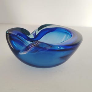 May include: A blue glass ashtray with a wavy, organic shape. The ashtray is made of thick glass and has a deep well.