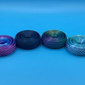 Puede incluir: Cuatro anillos giratorios de metal de diferentes colores con un borde exterior texturizado y un borde interior liso. Los anillos están dispuestos en una fila de izquierda a derecha, con los colores morado, negro, verde y azul.
