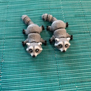 May include: Two gray and white raccoon figurines with black eyes and noses. The figurines are posed on their bellies with their tails curled up.