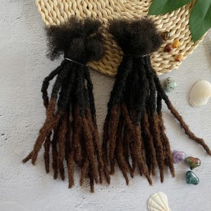 Peut inclure: Deux ensembles de dreadlocks en extensions de cheveux brun et noir. Les dreadlocks sont longues et épaisses, et elles sont disposées dans un style lâche et fluide.
