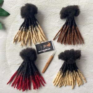 May include: Four sets of synthetic dreadlocks in various colors. The dreadlocks are black with blonde tips, black with red tips, brown with blonde tips, and black with brown tips. The dreadlocks are arranged on a white surface with a small card that says "Happy Locs Boutique" and a wooden needle.