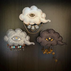 以下が含まれることがあります： 刺繍の目とビーズのアクセントが付いた、フェルト製の雲のブローチ3つ。雲は白、グレー、濃いグレーです。各ブローチには、クリア、ブルー、アンバーなど、異なる色のビーズが下部にぶら下がっています。