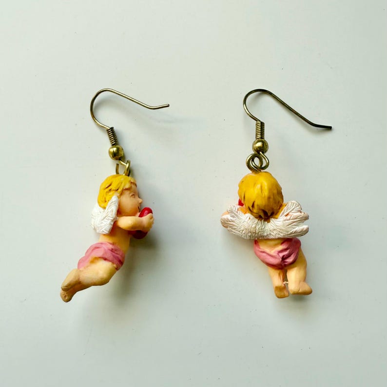 May include: A pair of earrings featuring cherub figures. Each cherub has blonde hair, white wings, and a pink garment, holding a red heart. The earrings have gold-colored hooks.