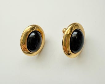 May include: A pair of gold-toned clip-on earrings. Each earring features a large, oval, black stone set within a gold frame. The earrings have a polished finish and a classic design.