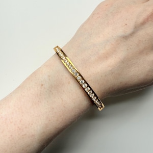 May include: A gold-colored bangle bracelet with a row of clear, sparkling gemstones. The bracelet is a simple, elegant design, perfect for everyday wear or special occasions. The gemstones are evenly spaced along the top of the bracelet.