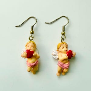 May include: A pair of dangle earrings featuring cherubic figures. Each earring depicts a small, smiling angel holding a red heart. The angels have blonde hair, white wings, and are wearing pink cloth. The earrings have gold-colored hooks.