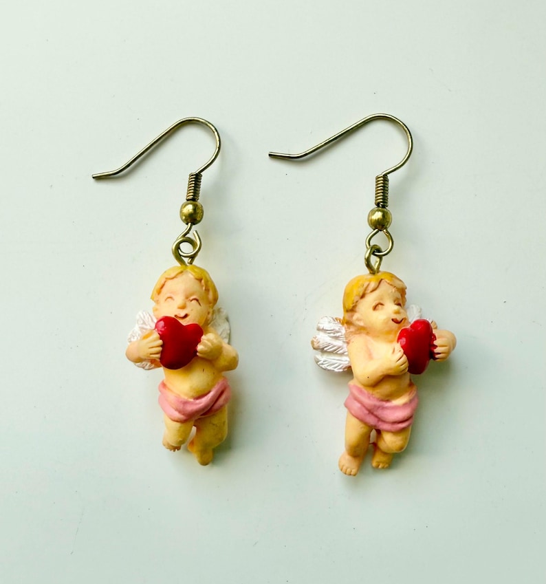 May include: A pair of dangle earrings featuring cherubic figures. Each earring depicts a small, smiling angel holding a red heart. The angels have blonde hair, white wings, and are wearing pink cloth. The earrings have gold-colored hooks.