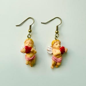 May include: A pair of earrings featuring cherubic figures holding red hearts. The cherubs have blonde hair, white wings, and are wearing pink cloth. The earrings have gold-colored hooks.