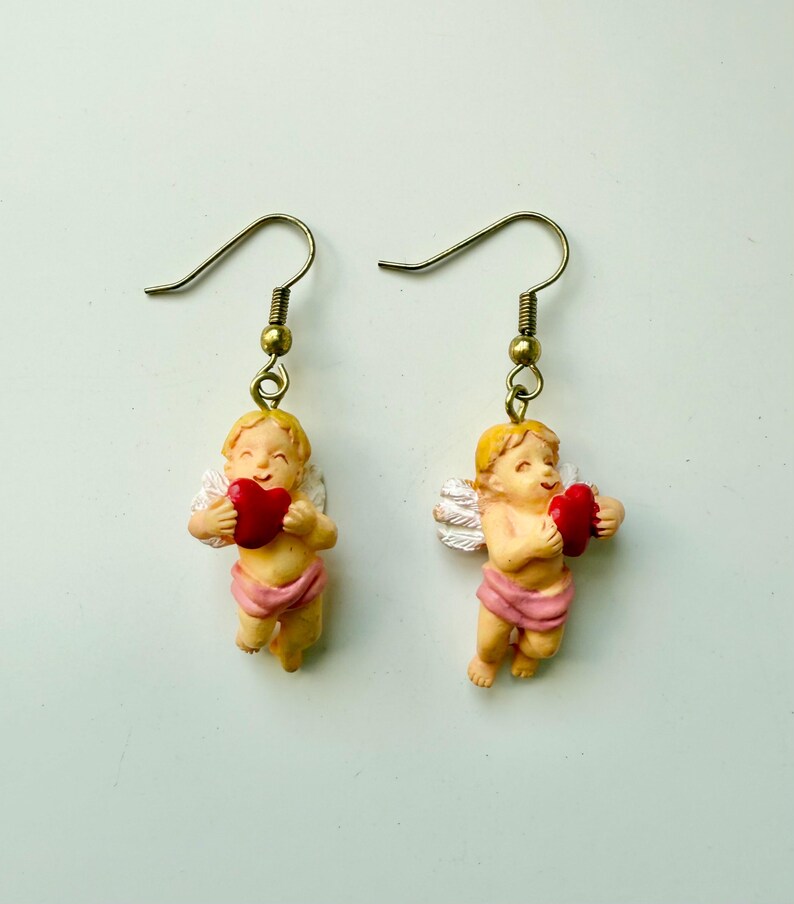 May include: A pair of earrings featuring cherubic figures holding red hearts. The cherubs have blonde hair, white wings, and are wearing pink cloth. The earrings have gold-colored hooks.