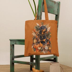 Könnte beinhalten: Eine rostorangefarbene Tragetasche mit einer grafischen Illustration eines Rottweilers, umgeben von Herbstlaub, Kürbissen, einer Laterne und einer Tasse mit dem Text Autumn Vibes. Das Design enthält warme Braun-, Orange- und Grüntöne.
