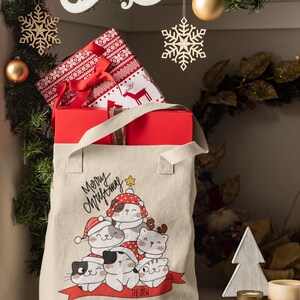 May include: A beige canvas tote bag featuring a graphic print of five cartoon cats wearing red and white holiday hats. The text Merry Christmas is printed above the cats, and the text The Crew is printed on a red banner below the cats.