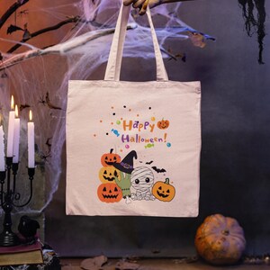 Könnte beinhalten: Eine beige Tragetasche aus Segeltuch mit einem bunten Grafikdruck mit dem Text Happy Halloween, drei orangefarbenen Kürbissen, einer kleinen Mumienfigur, einer grünen Monsterhand, einem lila Hexenhut, Fledermäusen und verstreuten Bonbons.