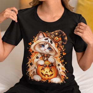 Könnte beinhalten: Ein schwarzes T-Shirt mit einer Grafik eines flauschigen Kätzchens, das eine geisterhafte Kapuze und einen Hexenhut mit Leopardenmuster trägt. Das Kätzchen hält einen leuchtenden orangefarbenen Kürbis, umgeben von orangefarbenen Farbspritzern und kleinen schwarzen Sternen.