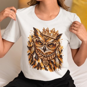 Könnte beinhalten: Ein weißes Kurzarm-T-Shirt mit einem Grafikdruck einer Eule, die eine goldene Krone, eine Brille mit Leopardenmuster und eine mehrlagige Halskette mit herzförmigem Anhänger trägt. Die Eule hat braune, schwarze und goldene Federn.