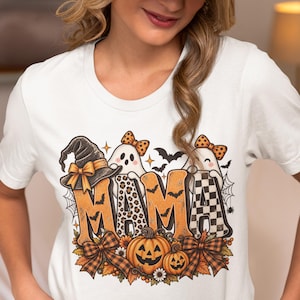 Könnte beinhalten: Ein weißes Kurzarm-T-Shirt mit einem Grafikdruck des Wortes MAMA in Orange, Leopardenmuster und schwarz-weißem Karomuster. Das Design zeigt zwei Geister mit orangefarbenen Schleifen, einen Hexenhut, Kürbisse und herbstliche Blätter.