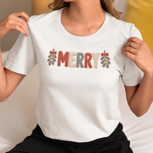 Könnte beinhalten: Ein weißes Kurzarm-T-Shirt mit dem Wort MERRY in einer texturierten, gesteppten Schriftart in abwechselnden Farben Rot, Creme und Blau. Der Schriftzug wird von kleinen, gestickten Illustrationen von Mistelzweigen mit roten Schleifen flankiert.