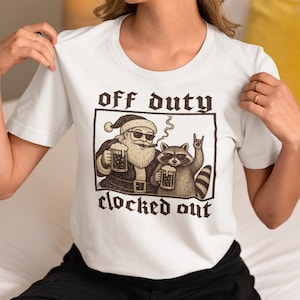 May include: A white short-sleeve t-shirt featuring a graphic print of Santa Claus and a raccoon wearing sunglasses, both holding beer mugs. Santa smokes a cigar, and the raccoon makes a hand gesture. The text reads off duty clocked out.