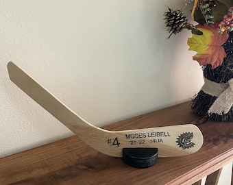 May include: A wooden hockey stick with a black puck base. The stick is engraved with the text "#4 MOSES LEIBELL '21-'22 14UA" and a logo.