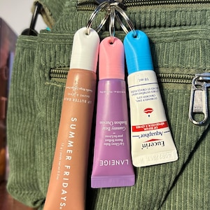 May include: Three lip balm tubes attached to a keyring. The tubes are pink, purple, and blue. The pink tube says "LANEIGE Gummy Bear Bonbon Ourson Lip Glowy Balm Baume Brillant pour les Lèvres". The purple tube says "SUMMER FRIDAYS. Lip Butter Balm". The blue tube says "Eucerin Aquaphor Reparateur a Levres Extremely Dry, Cracked Lips Vris Extremement Secs et Craquele 10 ml".