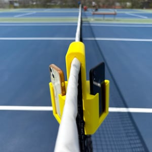May include: A yellow tennis net phone holder with two smartphones. One phone has a brown case, and the other is black. The holder is attached to the net post on a blue tennis court.