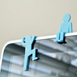 May include: Two light blue, stylised human figure-shaped clips. One figure is seated, the other is climbing. The clips are designed to attach to the edge of a device, such as a tablet or computer screen. The figures are made of a solid, smooth material.