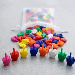 Puede incluir: Una colección de pequeñas figuras de mano de plástico de colores sólidos, cada una con el dedo medio extendido. El conjunto incluye varios colores como rosa, rojo, amarillo, azul, naranja, blanco y verde, con muchas saliendo de una bolsa de malla.