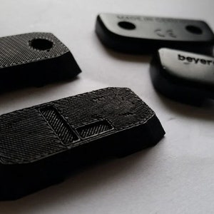 May include: A set of black plastic parts for headphones. The parts include two rectangular pieces with holes, a smaller rectangular piece with a groove, a round piece with the word "beyerdynamic" printed on it, and a smaller round piece with a groove.