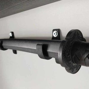 Könnte beinhalten: Ein schwarzes Katana-Schwert an einer Wand montiert. Das Schwert wird von zwei schwarzen Halterungen gehalten. Der Griff hat ein strukturiertes, gewebtes Muster. Die Klinge ist gebogen und hat eine Holzmaserung.