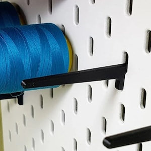 May include: A close-up of a white pegboard with two spools of turquoise thread and black plastic hooks. The thread is wound around the hooks.