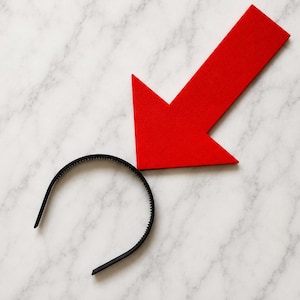 Puede incluir: Una diadema de plástico negro con una serie de dientes pequeños a lo largo del borde interior, colocada directamente debajo de una gran flecha roja sólida que apunta hacia abajo, hacia el centro de la diadema.