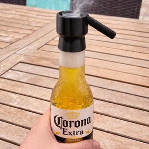 Puede incluir: Una botella de cerveza Corona Extra con un dispositivo negro en la parte superior, emitiendo vapor. La botella es sostenida por una persona. La botella de cerveza está sobre una mesa de madera, con una piscina en el fondo.