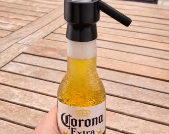 May include: A Corona Extra beer bottle with a black device on top, emitting vapour. The bottle is held by a person. The beer bottle is on a wooden table, with a pool in the background.