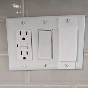 Puede incluir: Placa blanca de tomacorriente y interruptor de luz con un solo interruptor de luz y una sola toma de corriente. La placa tiene seis agujeros para tornillos y está montada en una pared de azulejos blancos.