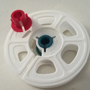 May include: White plastic film reel with a red plastic spindle and a blue plastic center. The reel has the text "REEL CORPORATION" printed on it.