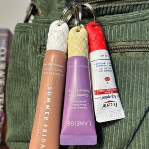 May include: Three lip balm tubes attached to a keychain. The tubes are pink, purple, and white with black text. The pink tube says "SUMMER FRIDA" and "Lip Butter Balm". The purple tube says "LANEIGE" and "Lip Glowy Balm". The white tube says "Eucerin Aquaphor Lip Repair".