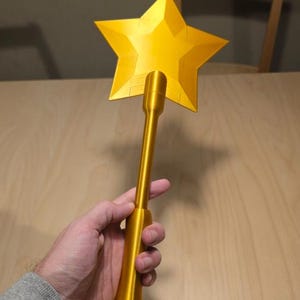 Puede incluir: Una varita dorada en forma de estrella con un mango largo. La varita se sostiene en la mano de una persona. La estrella tiene cinco puntas y está hecha de un material brillante y metálico. El mango es cilíndrico y también dorado.