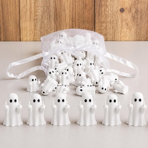 May include: A collection of small white ghost figurines with black oval eyes, each featuring raised arms with extended middle fingers. The figurines are arranged in a row in the foreground, with a larger group spilling out of a sheer white drawstring bag.