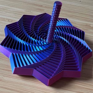 May include: A 3D-printed, geometric top in shades of purple and blue. The top has a star-like base with layered, angled segments that spiral inward. A cylindrical handle extends from the center. The top is on a wooden surface.
