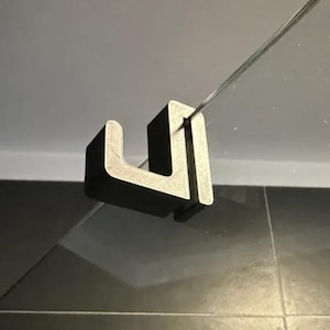 May include: A black, U-shaped bracket designed to hold a glass panel. The bracket is made of a solid material and is positioned to secure the glass. The background is a light grey and black tiled surface.