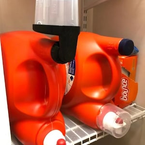 以下が含まれることがあります： オレンジ色の洗剤ボトル2本と透明な計量カップ。1本のボトルには白と赤の注ぎ口が付いています。ボトルは白いワイヤー棚にあります。背景にはBounceの乾燥機シートの箱が見えます。