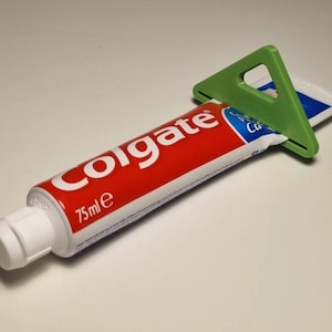 Puede incluir: Un tubo de pasta de dientes Colgate rojo y blanco con un exprimidor de plástico verde. El tubo está etiquetado con "75ml e". El tubo de pasta de dientes está parcialmente exprimido, con el exprimidor verde colocado en el extremo.