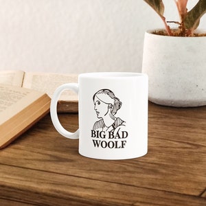 May include: White ceramic mug with a black line drawing of a woman's face and the text "BIG BAD WOOLF" on the front. The mug is sitting on a wooden table next to a book and a potted plant.