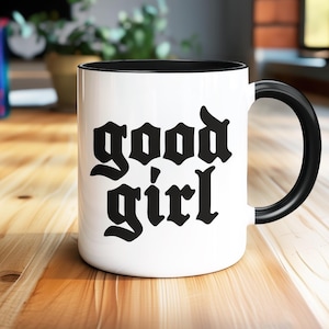 May include: White ceramic mug with a black interior and handle. The mug features the words "good girl" in a bold, gothic font. The mug is set on a wooden surface, with a blurred background including books and plants.