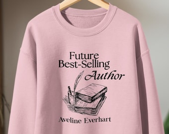 May include: A pale pink sweatshirt featuring the text "Future Best-Selling Author" and the name "Aveline Everhart" in black. A pen and ink drawing of books and a pen is below the text. The sweatshirt is suspended on a wooden hanger.
