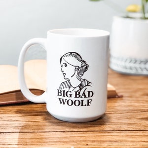 May include: White ceramic mug with a black and white illustration of a woman's face and the text "BIG BAD WOOLF". The mug is sitting on a wooden table next to a closed book.