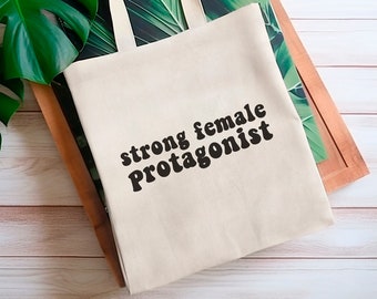May include: A white tote bag with black text that reads "strong female protagonist". The bag is sitting on a wooden surface with green leaves in the background.