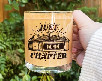 May include: A glass mug with a brown liquid inside. The mug has a black and white design of a book with the text "Just One More Chapter".
