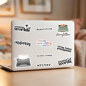 May include: A silver laptop covered in various stickers with text including weaver of worlds, #amwriting, storyteller, indoorsy, future best-selling author, strong female protagonist, too busy writing, writer, and weaver of words.